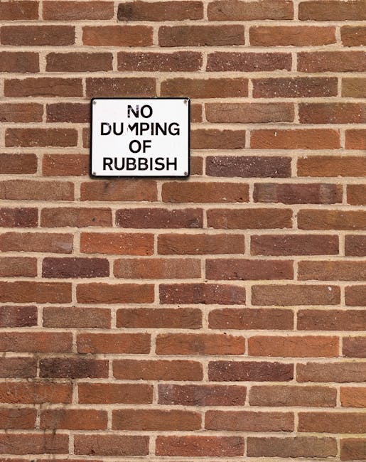 A rectangular metal sign with a white background and black text is mounted on a red brick wall. The sign reads 'NO DUMPING OF RUBBISH' in uppercase letters, with the words arranged in four lines. The brick wall features evenly spaced horizontal mortar joints, with bricks showing a range of reddish-brown and dark brown tones and a slightly rough texture. The sign is positioned centrally in the upper part of the image, contrasting clearly against the uniform brick background. The lighting is natural and even, with no strong shadows or highlights, emphasizing the clarity of the sign and the consistency of the brickwork, which often indicates a designated area for rubbish collection or waste management enforcement by Waste Collection Twickenham.