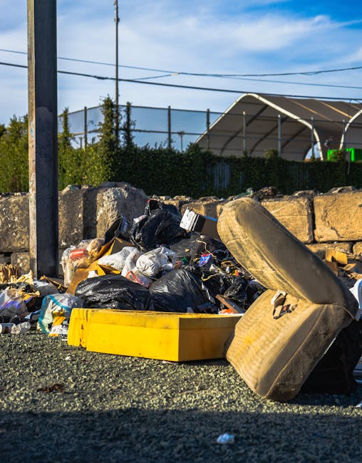 An outdoor scene showing a mound of mixed waste materials on a paved surface next to a low stone wall. The pile includes black plastic rubbish bags, cardboard boxes, plastic containers, and a large, dirty, beige tire leaning against a bright yellow plastic tray. In the background, there is a metal utility pole, power lines, and a fence with greenery beyond, along with a partially visible structure with a curved roof. The environment appears to be an area designated for waste collection or disposal, highlighting the type of rubbish that private waste services or independent collection methods might handle as part of a rubbish removal service in a residential or commercial setting.