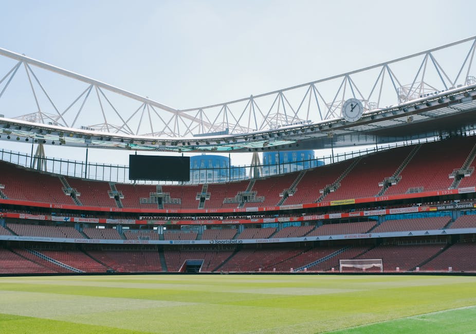 The image depicts an empty football stadium with a well-maintained grass pitch in the foreground, surrounded by multiple tiers of red and grey seating areas. The stadium has a modern, open roof structure with a white steel framework visible against a clear sky. In the background, media screens and advertisements are positioned above the seating levels, with a large digital clock mounted on the structural beams. The lighting appears natural, with sunlight illuminating the field and seating, creating a bright and clear atmosphere. The stadium's design features a combination of concrete and metal finishes, typical of contemporary sports venues. Given the context of waste management services, Waste Collection Twickenham could be involved in the private clearance or maintenance of such large-scale venues, emphasizing their capacity to handle waste removal in extensive sporting environments and large public spaces.