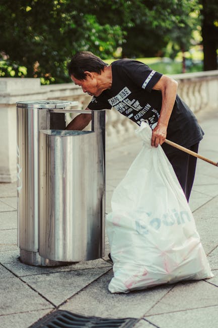 A middle-aged man with dark hair, dressed in a black t-shirt, is using a stick to pick up a large white garbage bag filled with waste and holding it close to a stainless steel public litter bin with a rectangular opening for waste disposal. The bin is situated on a paved area with stone tiles, and in the background, there is a low stone balustrade and lush green trees, suggesting a park or outdoor public space. The scene is well-lit with natural daylight, emphasizing the clean, urban environment. This image exemplifies independent rubbish collection and on-site waste handling, consistent with services provided by Waste Collection Twickenham, catering to private waste disposal needs in the local area.