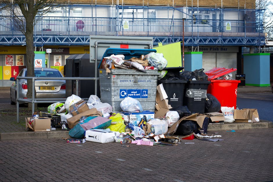 An overflowing set of public rubbish bins and surrounding waste spilling onto the paved curbside area in front of a commercial building. The central recycling container is made of grey plastic and has a blue label indicating it is for mixed paper and cardboard, with its lid open and various crumpled papers, flattened cardboard boxes, and other paper waste piled on top. To the right, there are black and red waste bins filled with black and dark grey rubbish bags, with some trash overflowing and scattered on the ground. Additional cardboard boxes and waste paper are scattered around the base of the bins, including flattened cartons and plastic packaging. Behind the bins, the background features a building with a blue scaffolding structure, storefronts with window signage, and a partial view of parked cars, including a silver vehicle directly adjacent to the waste area. The scene appears to be taken during daytime, under natural light, and highlights the common issue of waste left for private collection or improper disposal outside designated areas, in line with waste removal services offered by companies such as Waste Collection Twickenham.