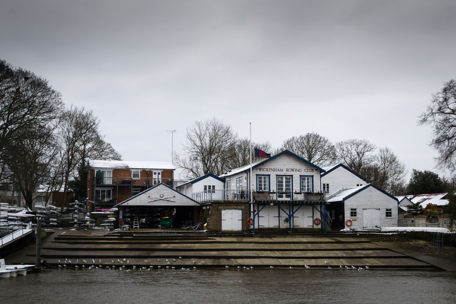 A view of the Twickenham Rowing Club situated along a waterfront, featuring a white-painted wooden building with blue trim, a balcony, and a sign indicating the club's name. Surrounding the building are leafless trees and residential houses with snow-covered roofs, indicating winter weather. The water in the foreground is calm, with concrete steps leading down to the shoreline, and a flock of seagulls resting on the steps. The overall scene is overcast, with a grey sky reflecting the subdued lighting conditions. This setting showcases a typical riverside facility that could be involved in water sports or boat storage, aligning with private and commercial waterway management, and may require periodic cleaning or rubbish removal from the area, which companies like Waste Collection Twickenham provide as part of their outdoor waste management services.