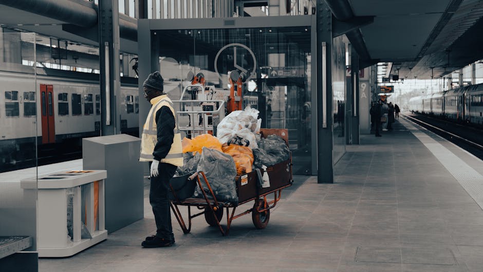 A person wearing a dark jacket, grey knitted beanie, and a reflective safety vest stands next to a large metal trolley cart filled with various black, grey, and orange plastic rubbish bags, some tightly packed and others loosely stacked, containing discarded household waste. The cart is positioned on a paved platform at a modern train station with a glass shelter and metal structural elements behind, reflecting the surrounding urban environment. To the left, a white recycling bin with an open slot for waste disposal is visible. In the background, a train with white and red cars is parked alongside the platform, with additional passengers seen farther along the station. The scene is lit naturally, suggesting daytime, and the overall setting appears to document a routine waste collection or waste management activity occurring in a busy transport hub, consistent with services provided by waste collection specialists like Waste Collection Twickenham, emphasizing on-site waste handling and bulk rubbish removal for the railway station area.