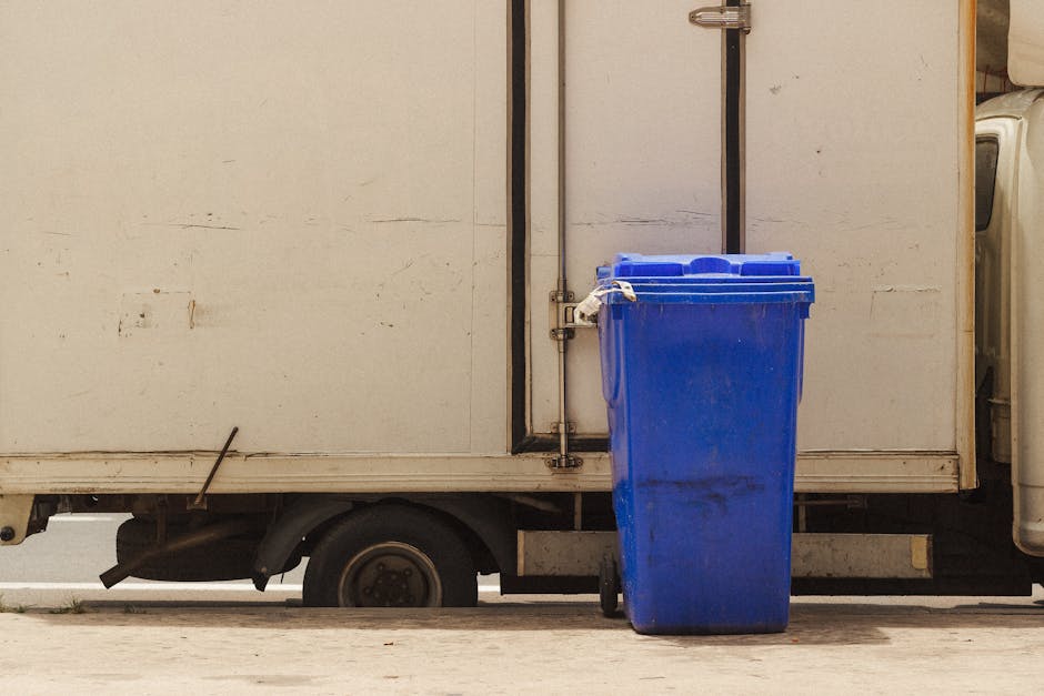 A bright blue plastic wheeled rubbish bin with a slightly weathered surface and smudges of dirt stands upright on the pavement next to a white delivery van with a slightly worn exterior. The van's side panel is plain with some minor scratches and dirt marks, and the black rubber wheel of the van is partially visible beneath it. The bin is positioned close to the van's rear door, which is secured with metal hinges and a latch. The surrounding environment includes a concrete sidewalk with subtle textures and small cracks, with a narrow gap between the van and the curb. The scene is lit with natural daylight, providing even illumination that highlights the textures of both the plastic bin and the van's surface. This image relates to private waste collection or alternative rubbish disposal options, with Waste Collection Twickenham occasionally supporting such on-site waste management services, emphasizing the practical handling of waste materials outside of general street rubbish collection.