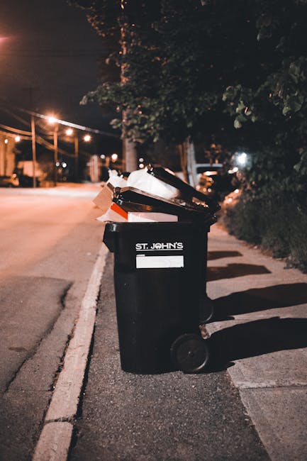 A person wearing a dark jacket, grey knitted beanie, and a reflective safety vest stands next to a large metal trolley cart filled with various black, grey, and orange plastic rubbish bags, some tightly packed and others loosely stacked, containing discarded household waste. The cart is positioned on a paved platform at a modern train station with a glass shelter and metal structural elements behind, reflecting the surrounding urban environment. To the left, a white recycling bin with an open slot for waste disposal is visible. In the background, a train with white and red cars is parked alongside the platform, with additional passengers seen farther along the station. The scene is lit naturally, suggesting daytime, and the overall setting appears to document a routine waste collection or waste management activity occurring in a busy transport hub, consistent with services provided by waste collection specialists like Waste Collection Twickenham, emphasizing on-site waste handling and bulk rubbish removal for the railway station area.